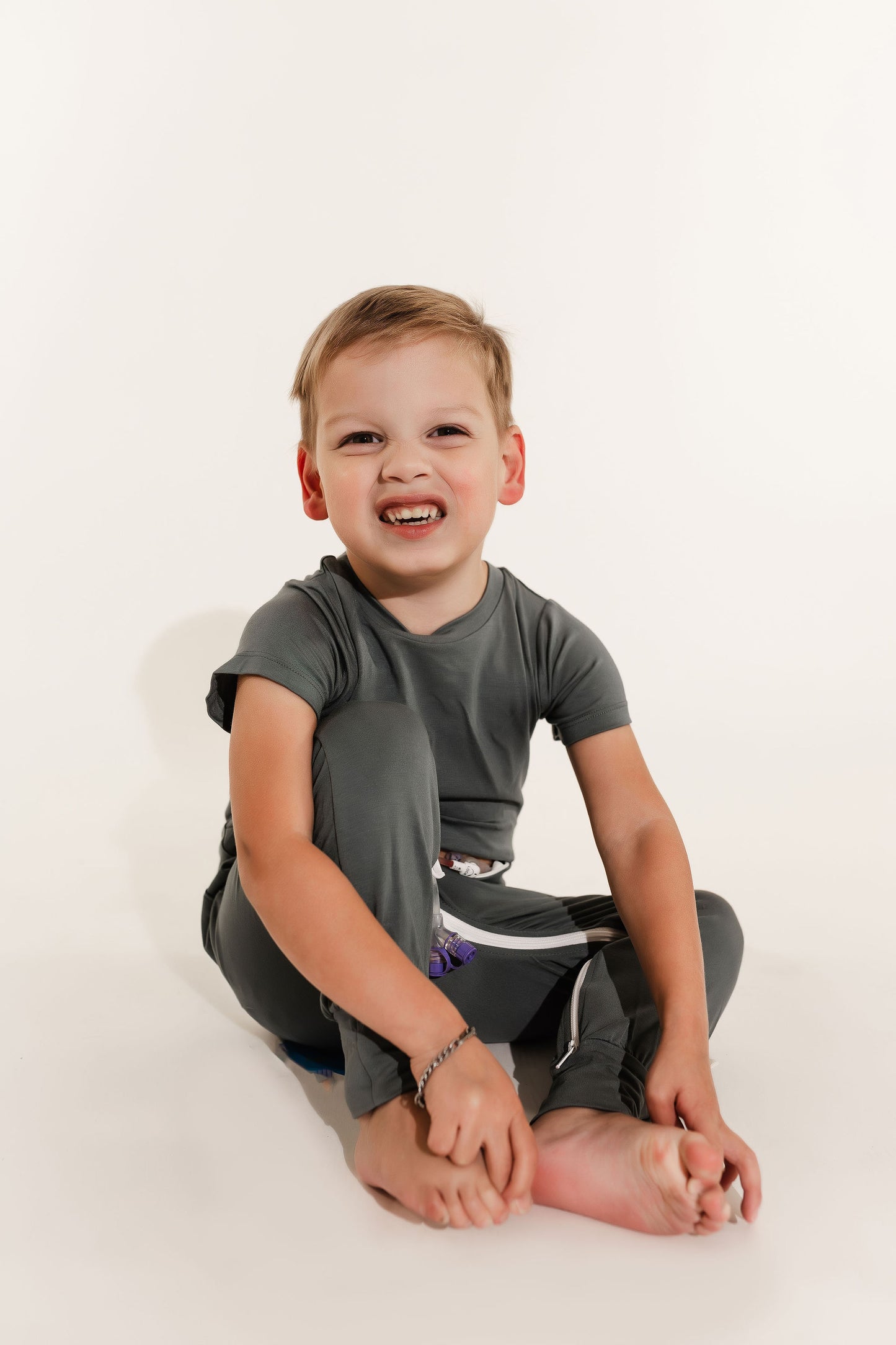 Child sitting on a white floor wearing a gray t-shirt and dark pants wearing a Bamboo Lyocell Zipease Romper g-tube gtube gastrostomy tube